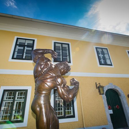 Eine bronzene Statue eines muskulösen Mannes, der seinen Arm anspannt, steht vor einem gelben Gebäude mit weißen Verkleidungen und mehreren Fenstern.