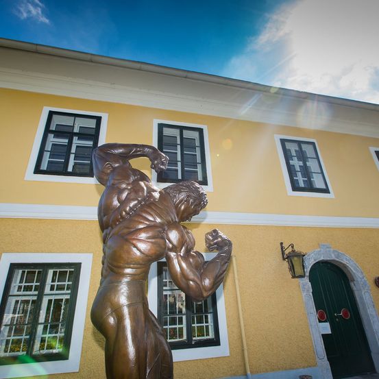 Eine bronzene Statue eines muskulösen Mannes, der seinen Arm anspannt, steht vor einem gelben Gebäude mit weißen Verkleidungen und mehreren Fenstern.