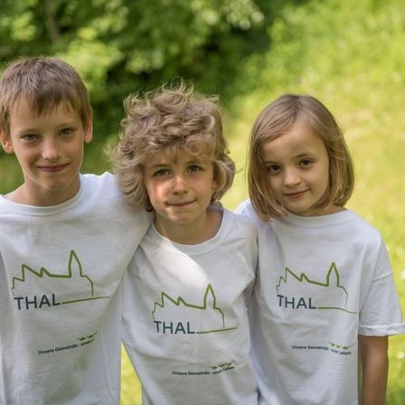 Drei Kinder in passenden weißen T-Shirts mit grünen Logos lächeln in einem Grasfeld. Der Junge links hat kurze Haare, während der Junge in der Mitte lockige Haare hat. Das Mädchen rechts hat lange Haare.