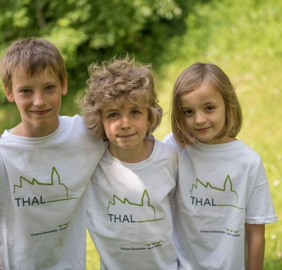 Drei Kinder in passenden weißen T-Shirts mit grünen Logos lächeln in einem Grasfeld. Der Junge links hat kurze Haare, während der Junge in der Mitte lockige Haare hat. Das Mädchen rechts hat lange Haare.