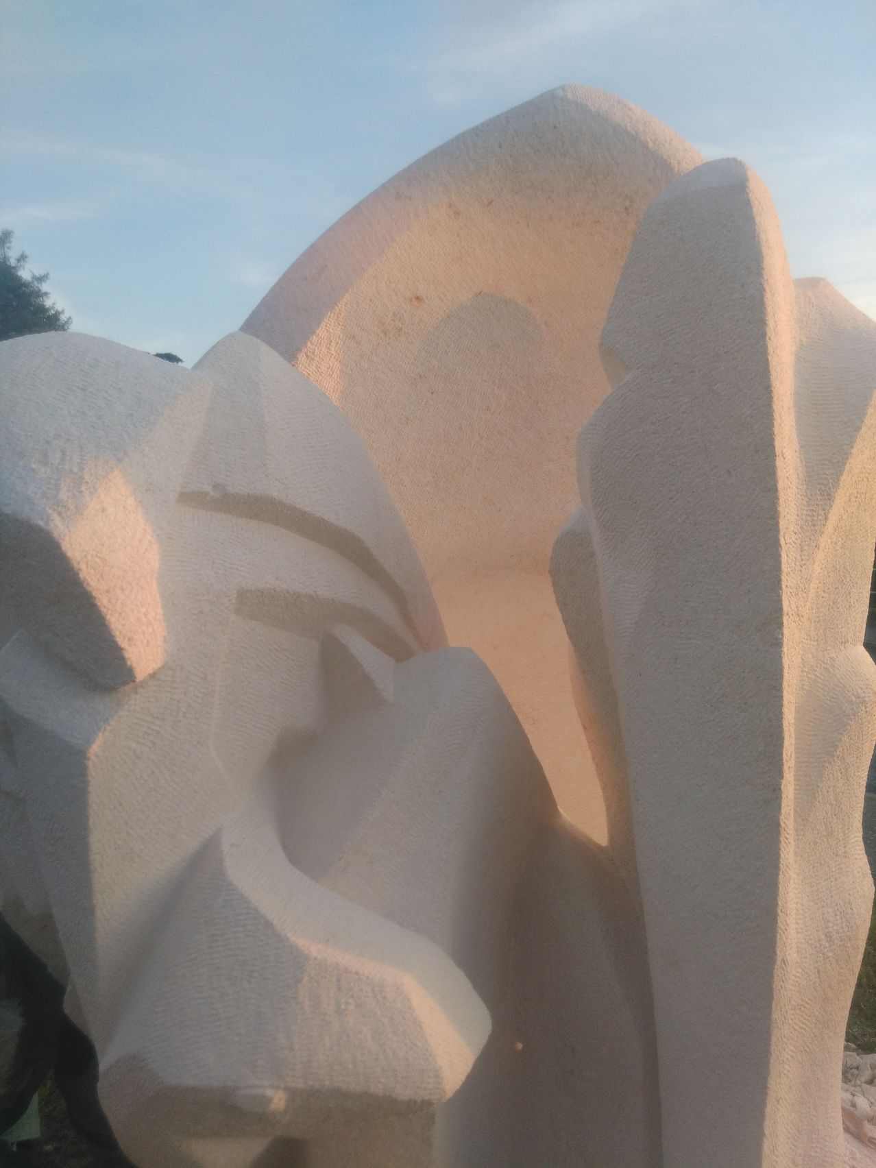 Eine große abstrakte Skulptur aus hellem Material, möglicherweise Stein oder Beton, mit einer strukturierten Oberfläche. Sie wird im Freien ausgestellt, und die Sonne wirft Schatten und Highlights auf die Skulptur.