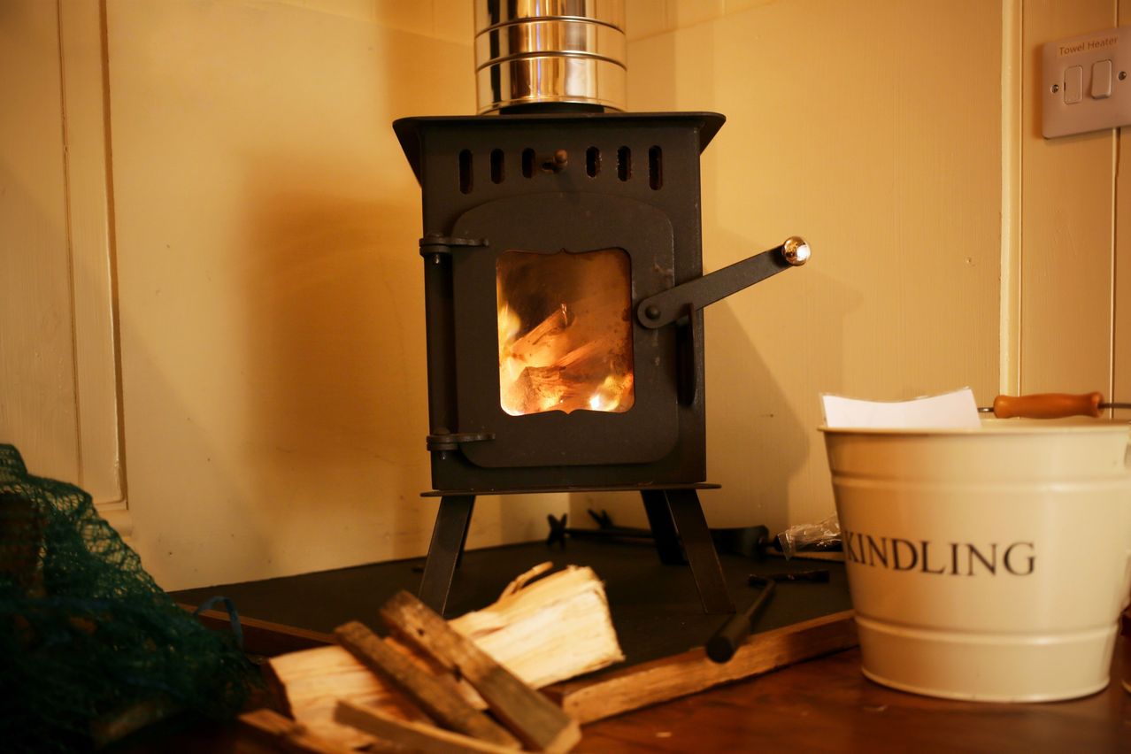Ein kleiner schwarzer Holzofen mit einem Metallschornstein auf einem Holztisch, ein Haufen Holz in der Nähe und ein weißer Eimer mit der Aufschrift 'KINDL'.