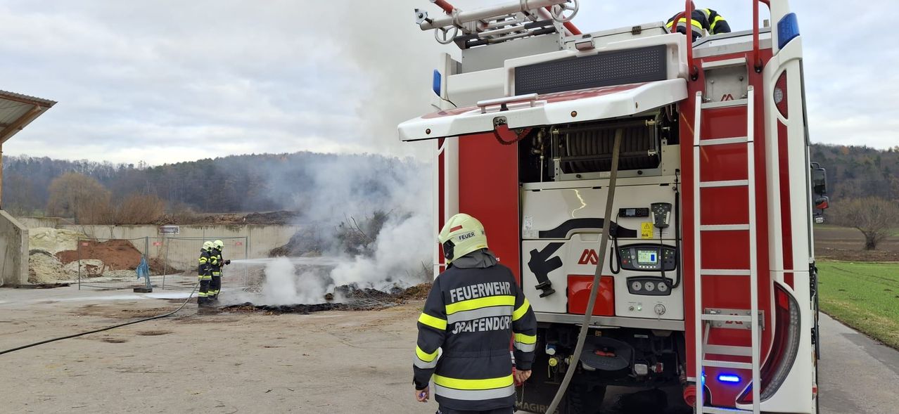 Ein Feuerwehrmann in einem gelben Helm steht neben einem Feuerwehrauto mit Rauch, der aus einem Brand im Hintergrund aufsteigt.