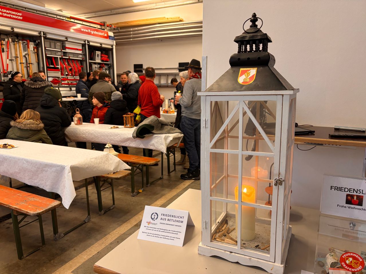 Eine Laterne mit einer brennenden Kerze steht auf einem Tisch, umgeben von Tischen und Menschen in einem Raum mit Feuerwehrausrüstung. Ein Schild befindet sich auf dem Tisch neben der Laterne.