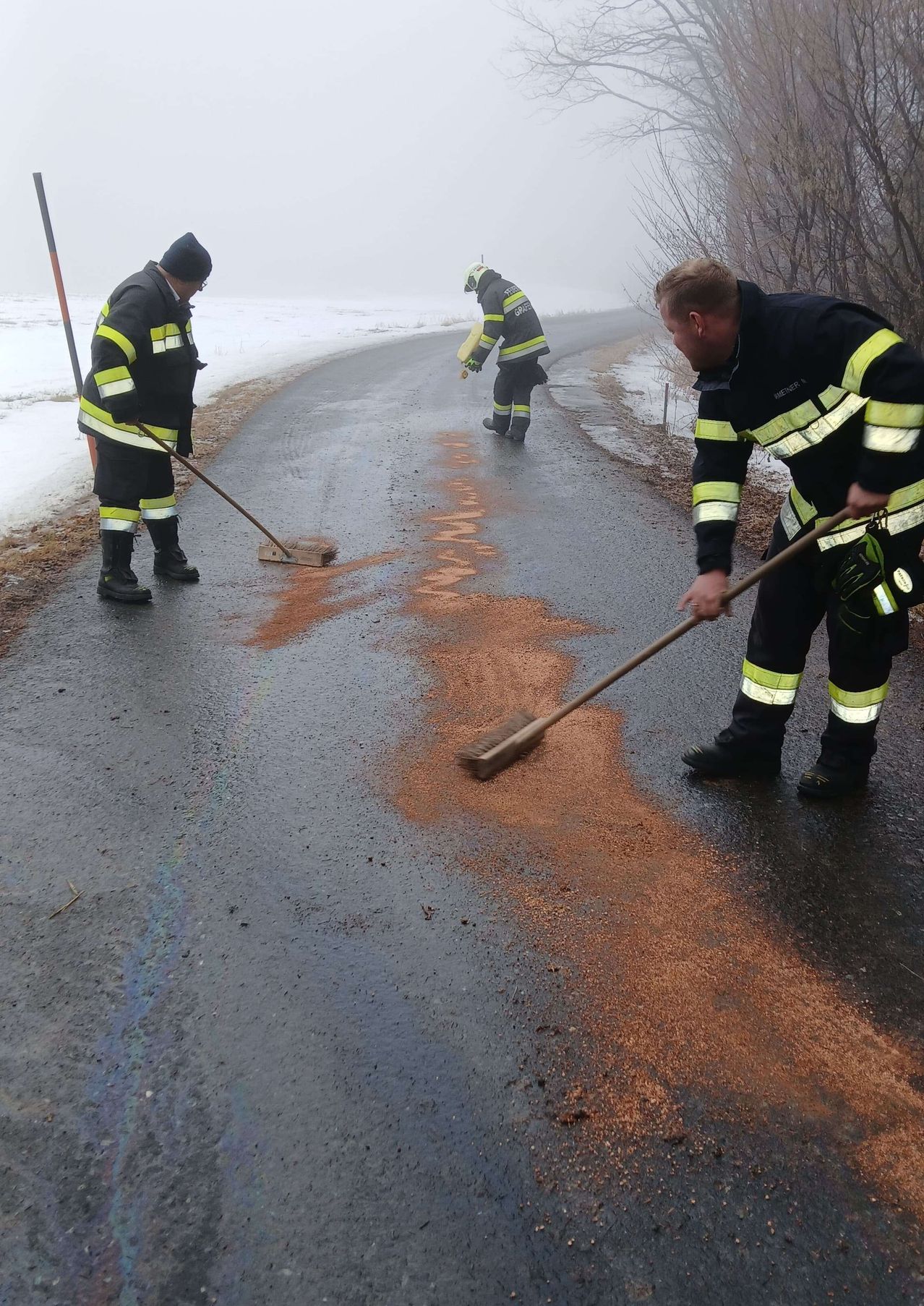 Drei Feuerwehrleute in Uniformen reinigen die Straße und benutzen Besen, um das braune Material aufzukehren. Die Straße hat einige Ölflecken und ist an den Seiten mit Schnee bedeckt.