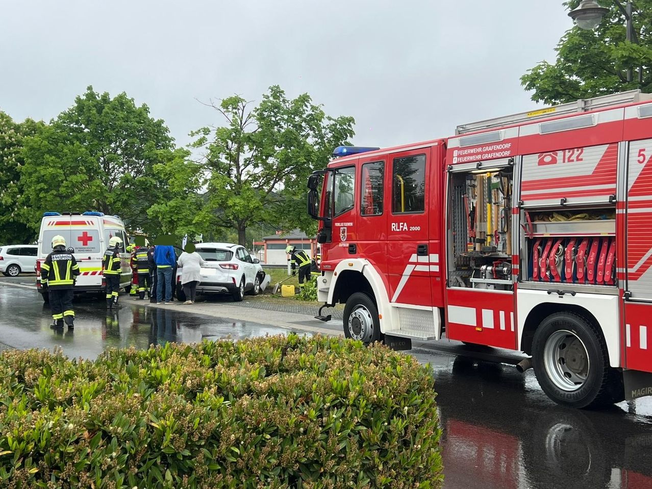 Feuerwehr und Rettungsdienst sind an Ort und Stelle in der Nähe eines beschädigten Autos und eines Busches bei nassem Wetter.
