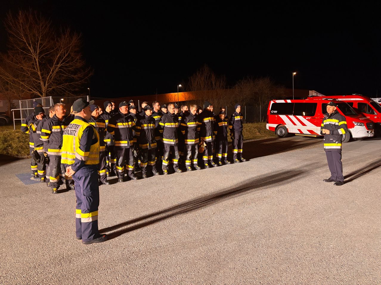 Eine Gruppe von Feuerwehrleuten steht bei Nacht in einer Reihe. Sie tragen reflektierende Kleidung. Ein roter Feuerwehrwagen ist in der Nähe geparkt.