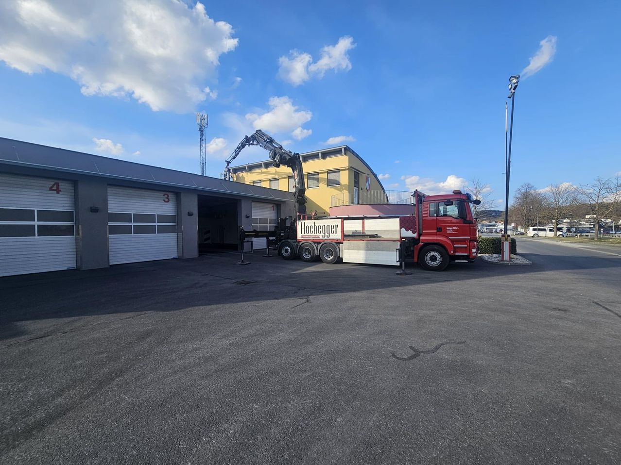 Ein roter Lkw mit einem Kran steht auf einem Parkplatz vor einer Garage mit der Nummer 3. In der Ferne befinden sich Bäume und ein Turm.