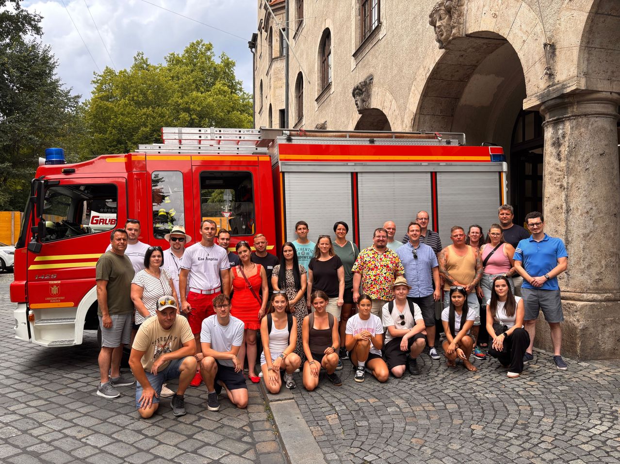 Eine Gruppe von Menschen posiert für ein Foto vor einem roten Feuerwehrauto, mit einem Gebäude und einem Baum im Hintergrund.