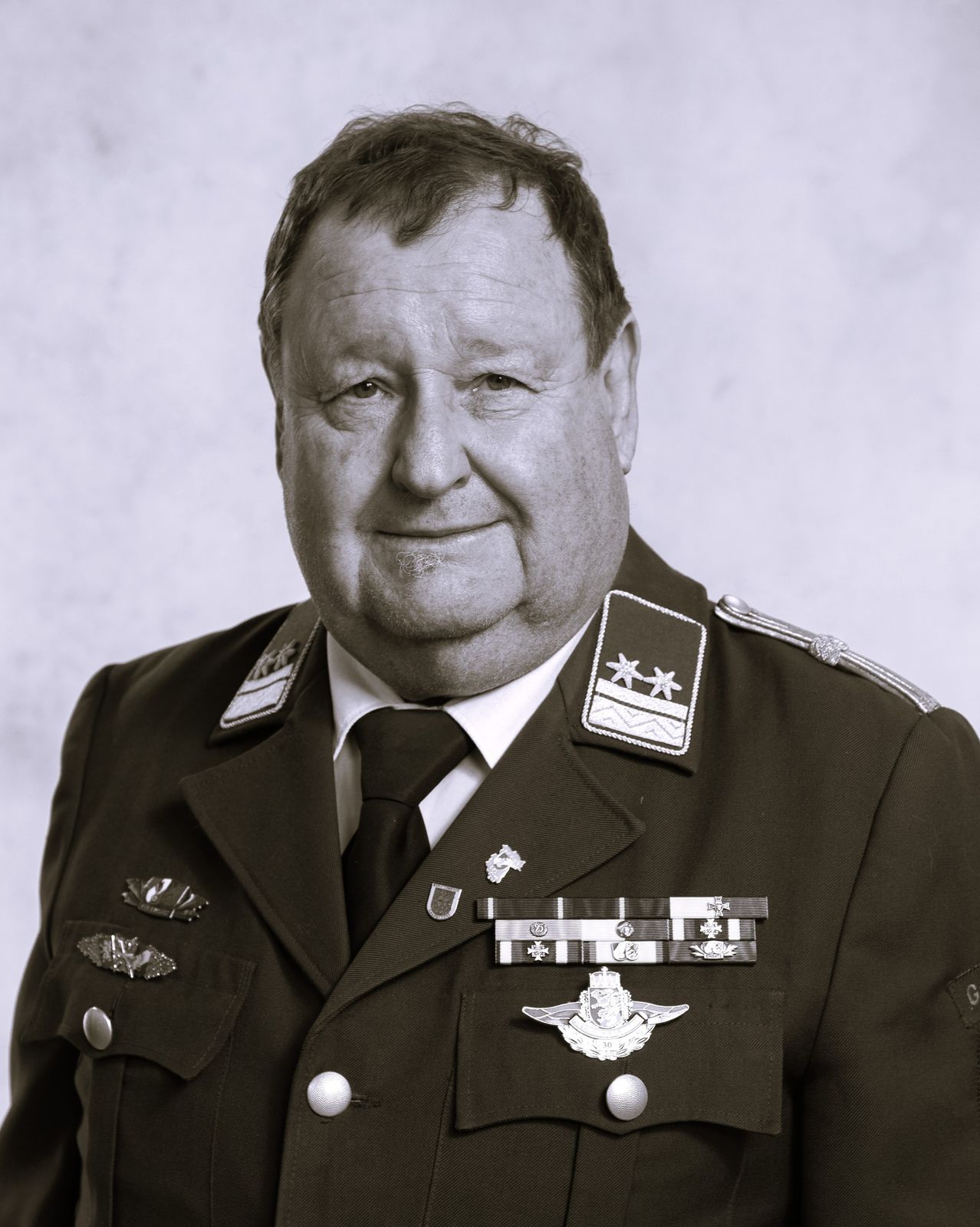 A black-and-white portrait of a man in a military uniform, smiling and looking directly at the camera. He wears a tie and several medals on his chest.