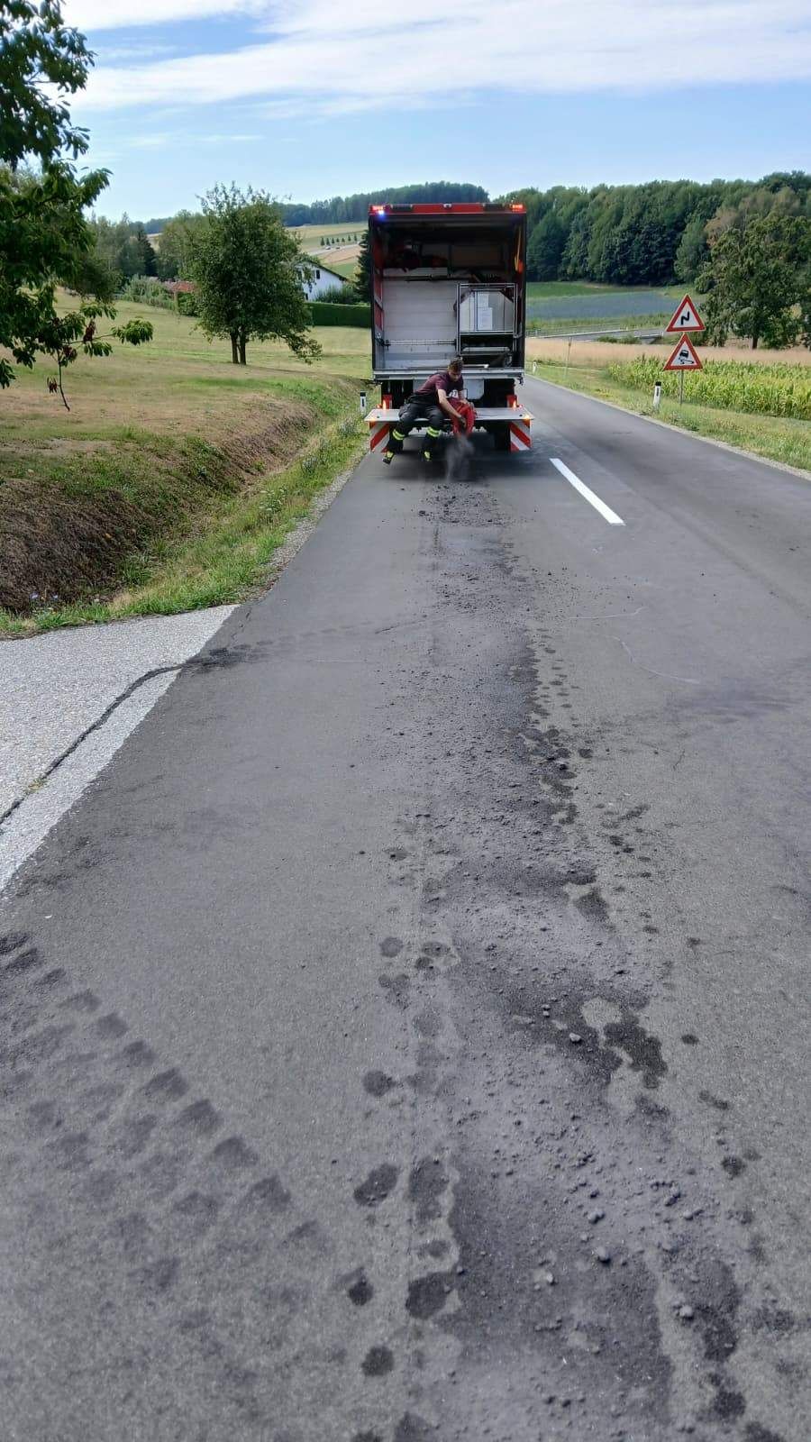 Ein Arbeiter in einer reflektierenden Weste trägt eine Substanz auf die Straße auf und hinterlässt Spuren auf dem Asphalt. Die Straße ist von Gras und Bäumen umgeben.