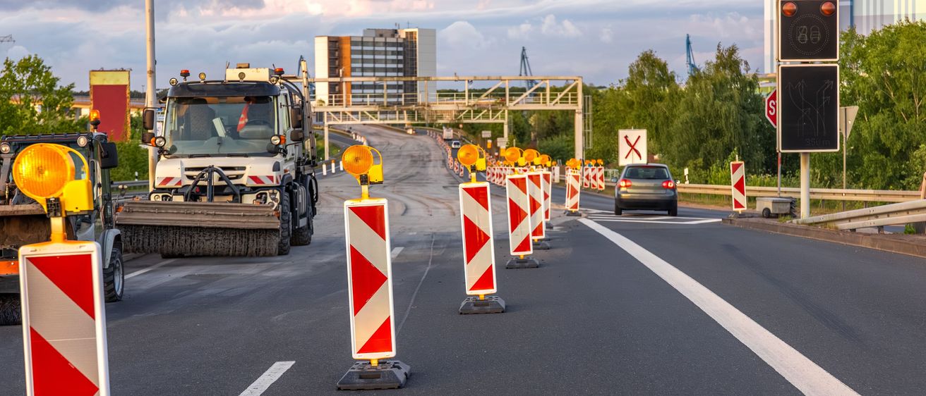Eine Autobahn mit einem Straßenbaufahrzeug, Verkehrskegel und einem Auto in der Ferne. Im Hintergrund sind Gebäude zu sehen.