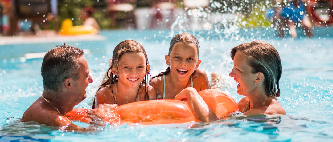 Eine vierköpfige Familie genießt einen sonnigen Tag am Pool, wobei zwei junge Mädchen einen orangefarbenen Schwimmflügel halten und lächeln.