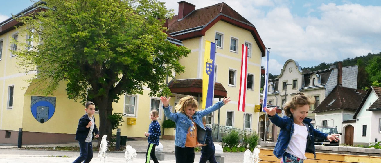 Kinder spielen und springen in einem Brunnen vor einem gelben Gebäude mit Fahnen auf einem Stadtplatz.