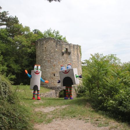 Zwei Maskottchen in bunten Kostümen stehen vor einem alten Steinturm in einem Park.