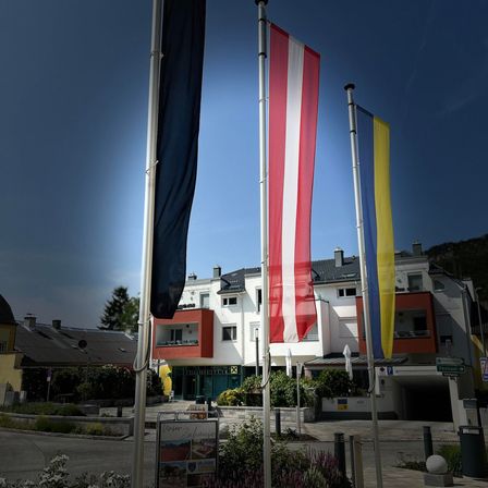Einige Fahnen wehen vor einem weiß gestrichenen Gebäude mit roten Akzenten, umgeben von Grün, unter einem blauen Himmel.