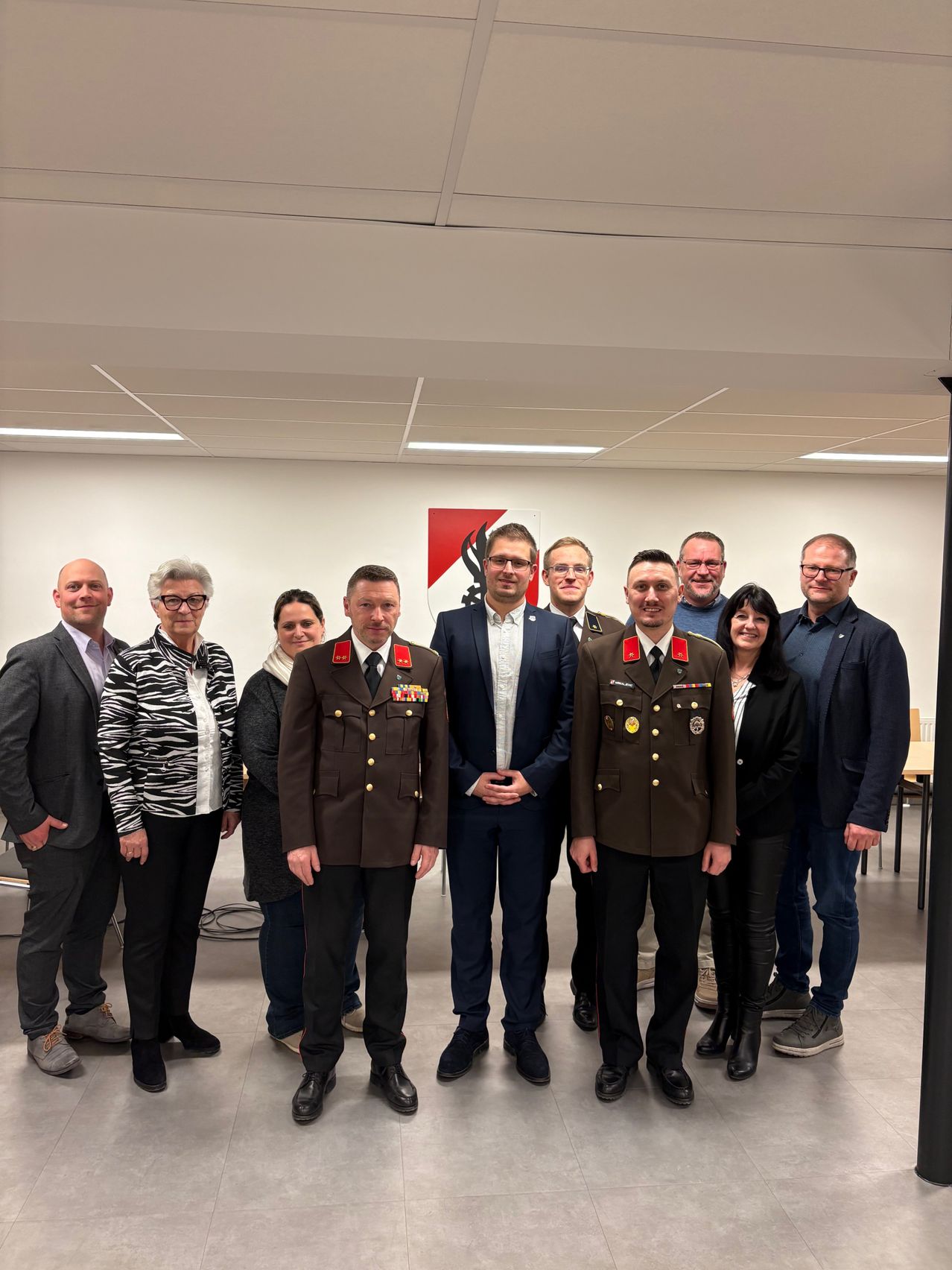 Eine Gruppe von Personen, einige in Militäruniformen und andere in formeller Kleidung, stehen zusammen in einem Raum mit einem roten Schild im Hintergrund.