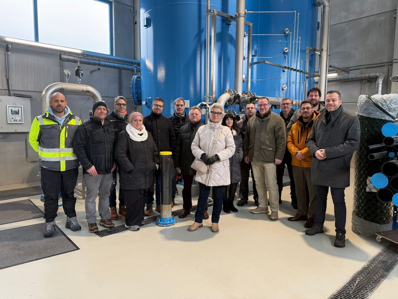 Eine Gruppe von Menschen steht in einer Fabrik mit blauen Tanks. Sie tragen Winterkleidung und scheinen für ein Foto zu posieren.