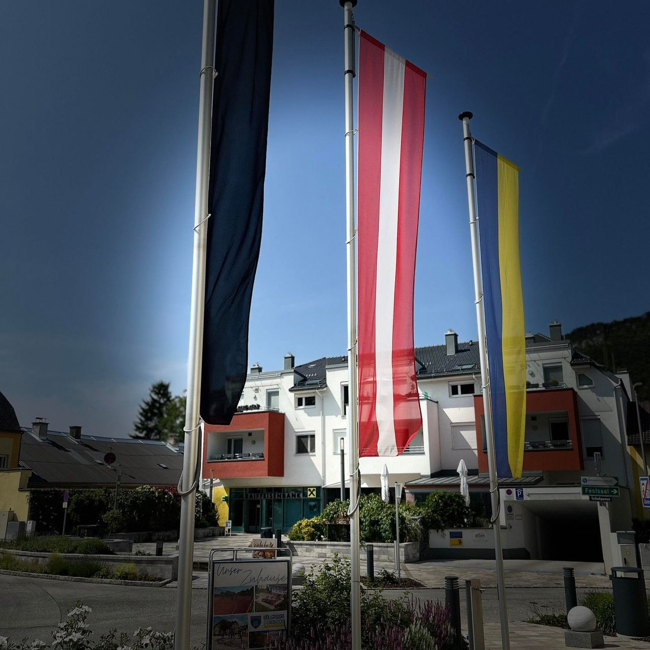 Drei Fahnen sind vor einem Gebäude mit einer grünen Tür auf Stangen angebracht. Die Fahnen sind in verschiedenen Farben, wobei eine schwarz ist. Das Gebäude hat mehrere Fenster und ist von Pflanzen und Bäumen umgeben.