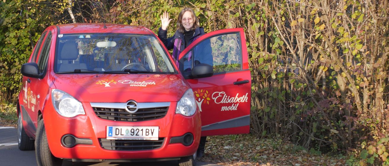 Eine Frau steht neben einem roten Opel-Auto mit offener Tür und winkt. Das Auto hat das Kennzeichen DL 921 BV. Hinter ihr stehen Bäume mit grünen Blättern.