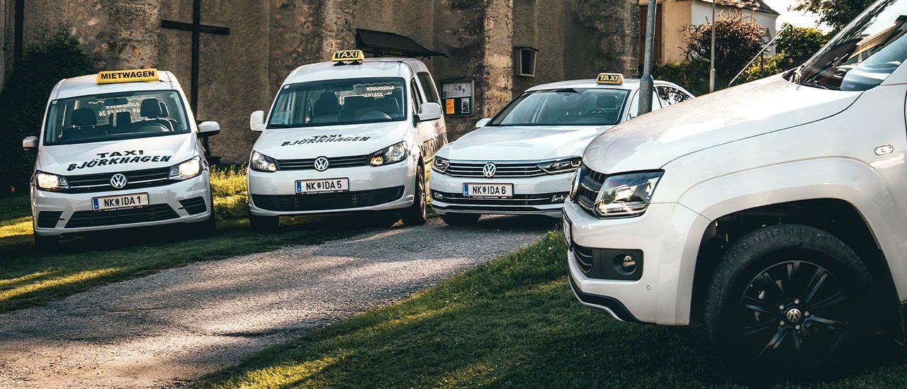 Mehrere weiße Taxis sind vor einem alten Gebäude mit Bogenfenstern und einem Kreuzsymbol am Straßenrand geparkt.