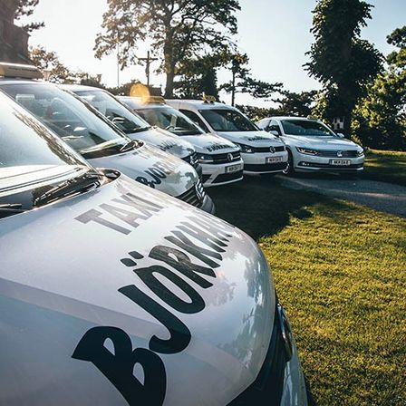 Mehrere Autos sind auf einem Rasenbereich geparkt, mit Bäumen im Hintergrund. Die Autos haben das Wort Bjorkman an ihren Seiten.