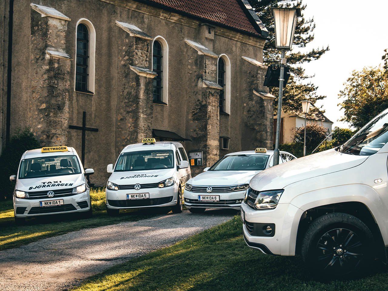 Vier weiße Taxis mit gelben Schildern sind vor einer Kirche geparkt. Bäume und Straßenlampen sind im Hintergrund zu sehen.