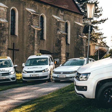 Vier weiße Taxis mit gelben Schildern sind vor einer Kirche geparkt. Bäume und Straßenlampen sind im Hintergrund zu sehen.