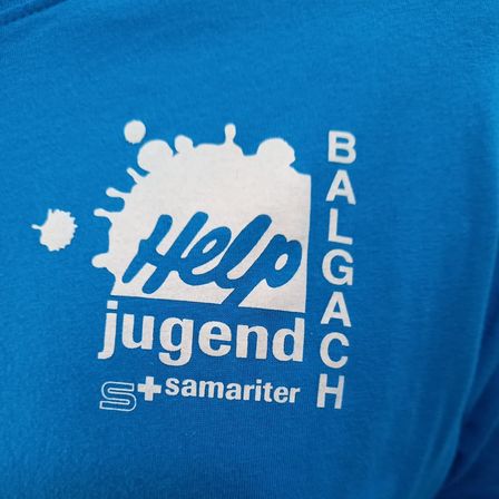Ein blaues T-Shirt mit einem weißen Logo, das die Wörter 'Hilfe' und 'Jugend' enthält. Das Logo umfasst 'BALGAC' und 'S+samariter'. Das Shirt ist für Jugendliche konzipiert.
