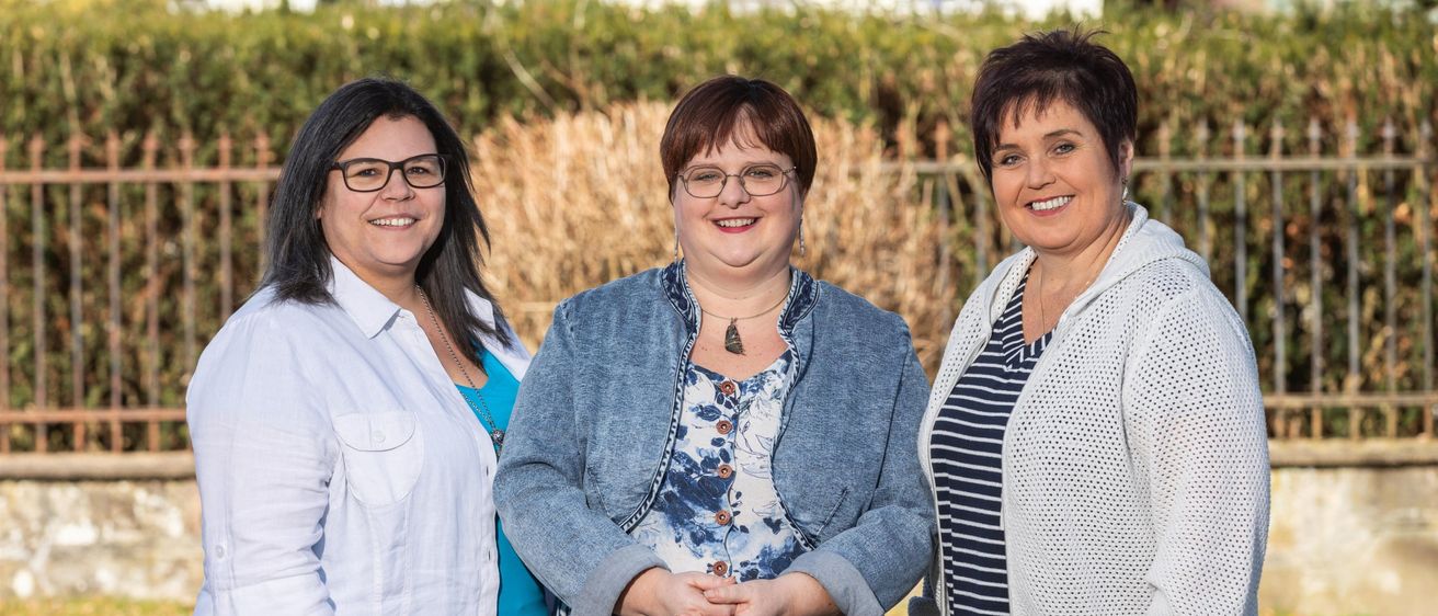 Three women stand together in a garden. They are smiling and appear to be posing for a photograph. Each woman wears a different style of clothing.