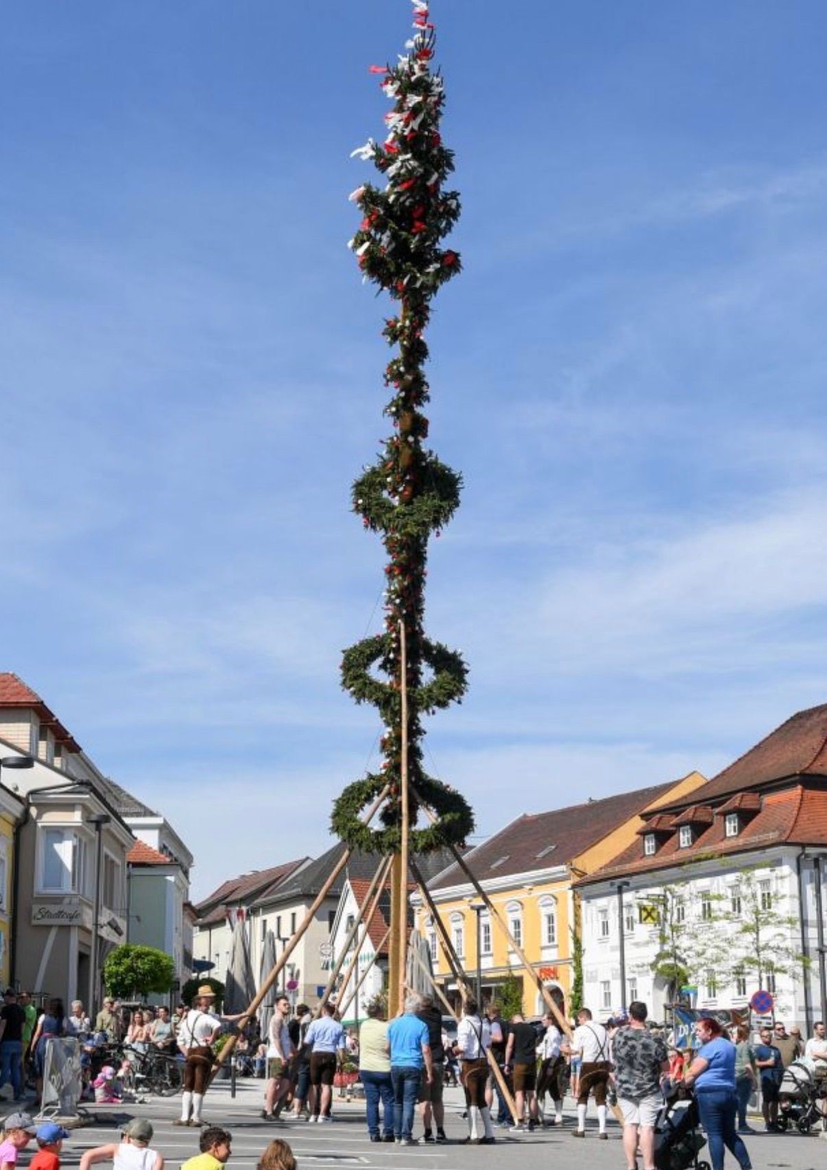 Ein hoher Weihnachtsbaum in der Mitte einer Straße, umgeben von Gebäuden mit verschiedenen architektonischen Designs, unter einem klaren blauen Himmel.