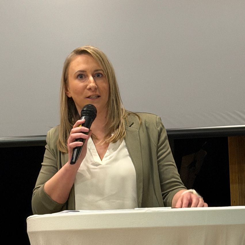 Eine Frau spricht in ein Mikrofon am Rednerpult, trägt eine grüne Jacke über einem weißen Hemd. Sie scheint ein Publikum anzusprechen, mit einer leeren Projektionsleinwand hinter ihr.