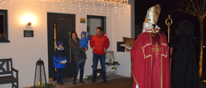 Eine vierköpfige Familie steht vor einem Haus mit einem Priester in festlicher Kleidung. Die Familie besteht aus einer Frau, die ein Kind hält, einem Mann und zwei Kindern. Das Haus hat eine Tür und ein Fenster mit einer Lampe.