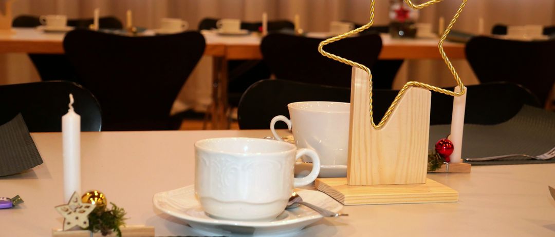 Ein festlicher Esstisch ist mit weißen Tassen, Untertassen und einer goldenen Sternmitte dekoriert. Der Tisch ist von schwarzen Stühlen und einer weißen Tischdecke umgeben.