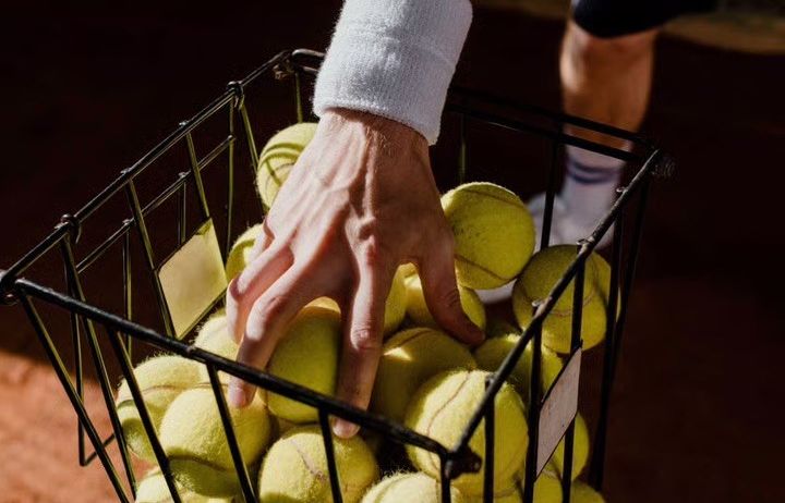 Die Hand eines Tennisspielers hält einen Korb voller Tennisbälle auf einem Tennisplatz.