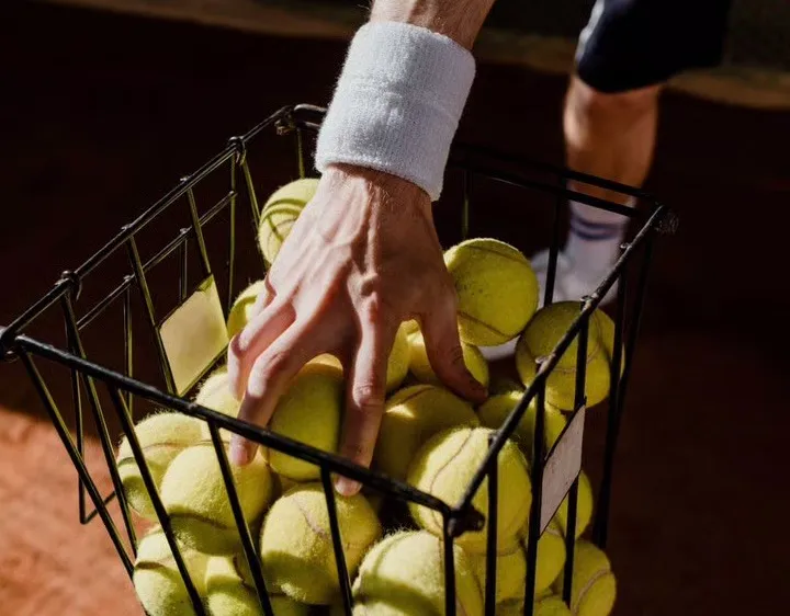 Die Hand eines Tennisspielers hält einen Korb voller Tennisbälle auf einem Tennisplatz.