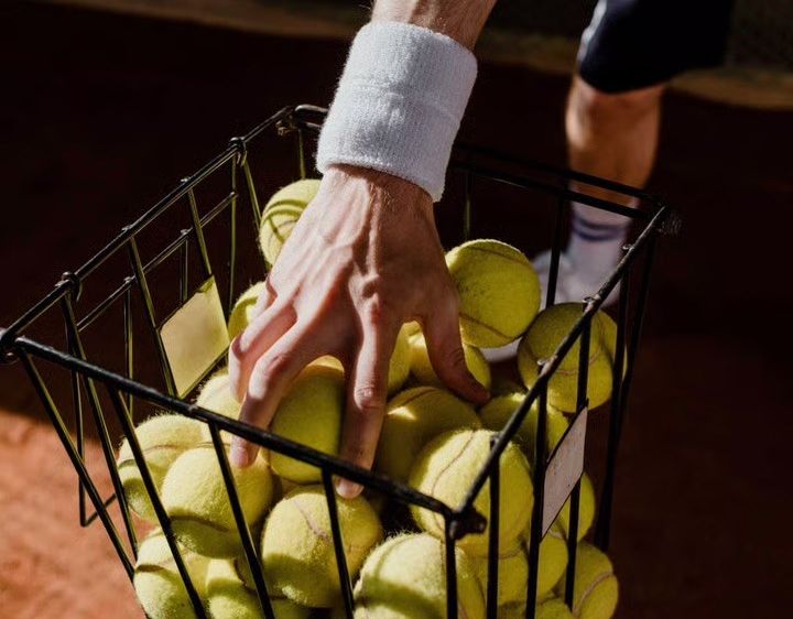 Die Hand eines Tennisspielers hält einen Korb voller Tennisbälle auf einem Tennisplatz.