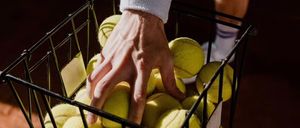 A tennis player's hand holds a basket full of tennis balls on a tennis court.