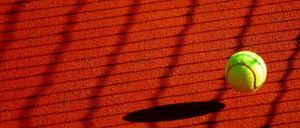A tennis ball sits on a red tennis court, its shadow cast on the clay ground. The court's texture is visible, with red clay and white lines.