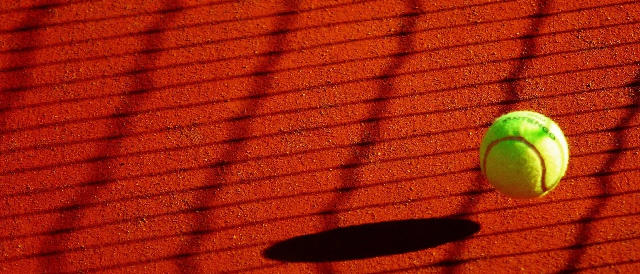 Ein Tennisball liegt auf einem roten Tennisplatz, sein Schatten fällt auf den Lehmboden. Die Textur des Platzes ist sichtbar, mit rotem Lehm und weißen Linien.