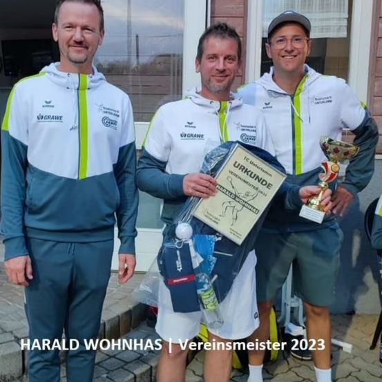 Drei Männer stehen zusammen mit Trophäen, einer hält ein Zertifikat. Sie tragen weiße und grüne Uniformen mit 'GRAWE' auf der Vorderseite. Der Mann auf der linken Seite hat einen Bart, und der Mann in der Mitte lächelt. Der Mann auf der rechten Seite trägt eine Brille und einen Hut.