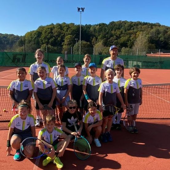 Eine Gruppe junger Tennisspieler posiert für ein Foto auf einem Tennisplatz, trägt einheitliche Kleidung und hält Schläger. Im Hintergrund befindet sich ein grüner Zaun mit einem Schild und einem Gebäude.