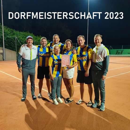 Eine Gruppe von Personen, möglicherweise ein Sportteam, steht auf einem Tennisplatz mit einem Schild, das Dorfmeisterschaft 2023 besagt. Sie sind in Sportbekleidung gekleidet.