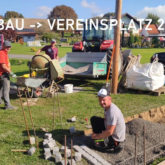Drei Männer arbeiten auf einer Baustelle, einer kniet und einer steht. Geräte wie ein Schubkarren, ein Zementmischer und ein Traktor sind vorhanden. Ein Haus und Bäume sind im Hintergrund zu sehen.