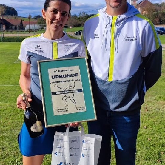 Ein lächelnder Mann und eine Frau stehen auf einem Feld mit einem gerahmten Zertifikat und einer Flasche Champagner. Die Frau trägt einen blauen Rock.