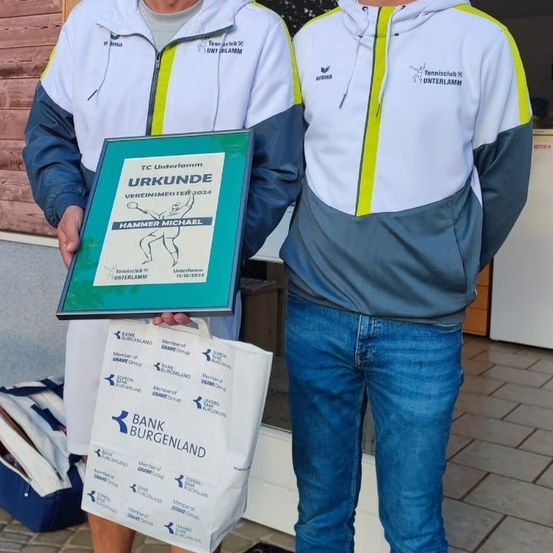 Zwei Männer stehen zusammen, einer hält ein gerahmtes Zertifikat und der andere eine weiße Tasche. Sie tragen passende weiße und blaue Jacken mit gelben Akzenten. Die Tasche steht 'Bank Burgenland'. Das Zertifikat steht 'TC Unterlamm' und 'URKUNDE' mit einer Zeichnung einer Person.