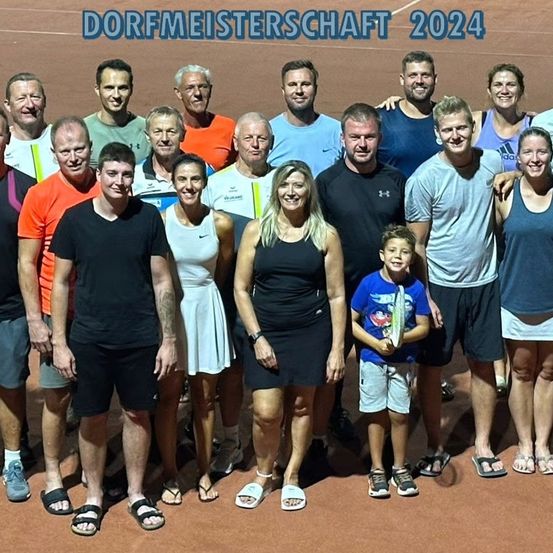 Eine Gruppenaufnahme von Erwachsenen und Kindern bei einer Sportveranstaltung im Jahr 2024. Sie stehen auf einem Tennisplatz und tragen Freizeitkleidung. Ein Junge hält einen Tennisschläger.