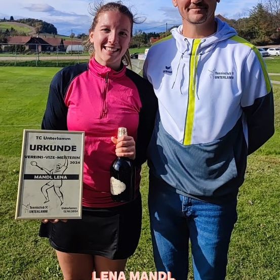 Eine Frau und ein Mann stehen auf einem Feld und halten eine Trophäe und eine Flasche. Die Frau lächelt und der Mann hält eine Flasche. Sie tragen beide Sporttrikots.