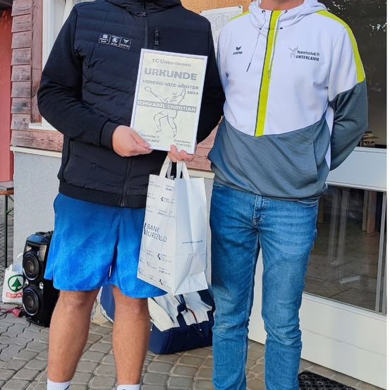 Zwei Männer stehen draußen, einer hält ein Zertifikat und einen Beutel. Einer trägt eine schwarze Jacke und blaue Shorts, der andere eine weiße Jacke mit gelbem Besatz. Hinter ihnen steht ein Gebäude und ein Lautsprecher.