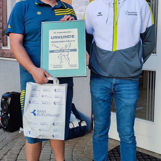 Zwei Männer stehen zusammen und halten ein gerahmtes Zertifikat in der Hand, einer trägt ein blaues Poloshirt und der andere eine weiße Kapuzenjacke mit grauen Akzenten. Sie stehen vor einem Gebäude mit einer Glastür.